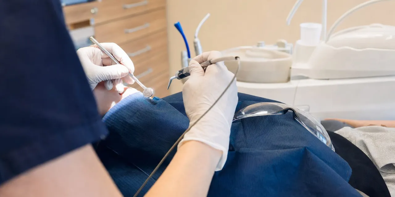 Zahnmedizinische Fachkraft in dunkelblauer Kleidung und weißen Handschuhen behandelt einen Patienten mit Spiegel und Instrument im Behandlungszimmer.